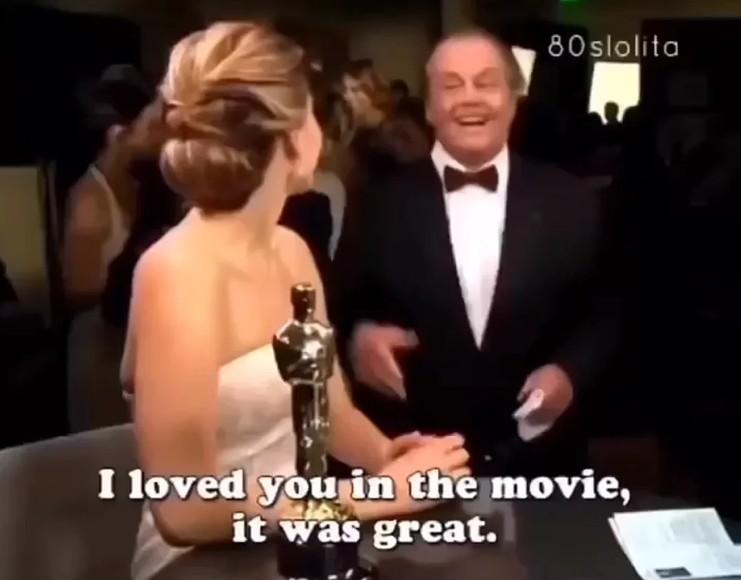 A woman in a white dress holding an Oscar statue talks to a smiling man in a tuxedo. The caption reads, "I loved you in the movie, it was great." A crowd and lights are visible in the background.