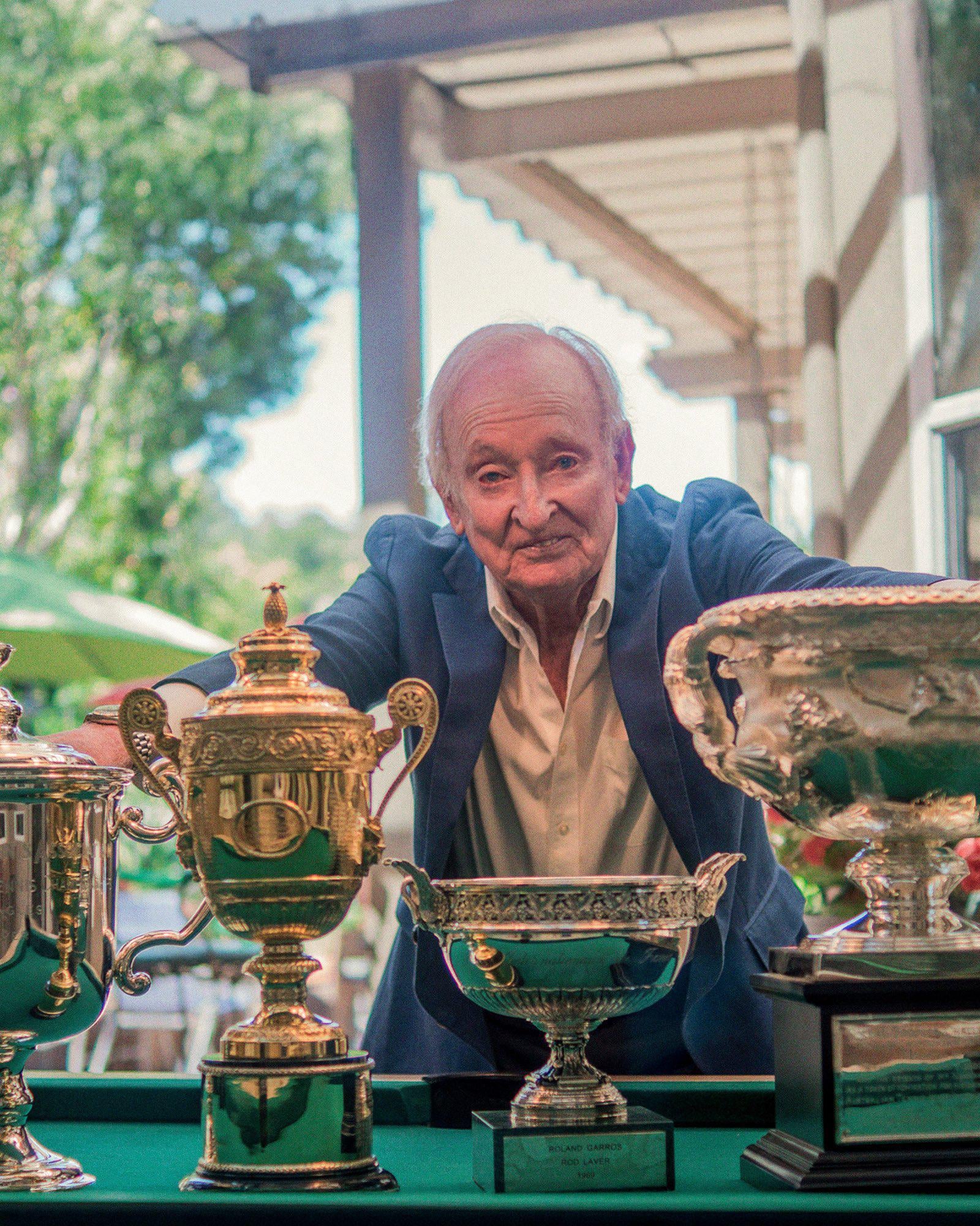 An older man in a blue blazer stands behind a table displaying several large, ornate trophies outdoors, with greenery and a building visible in the background.