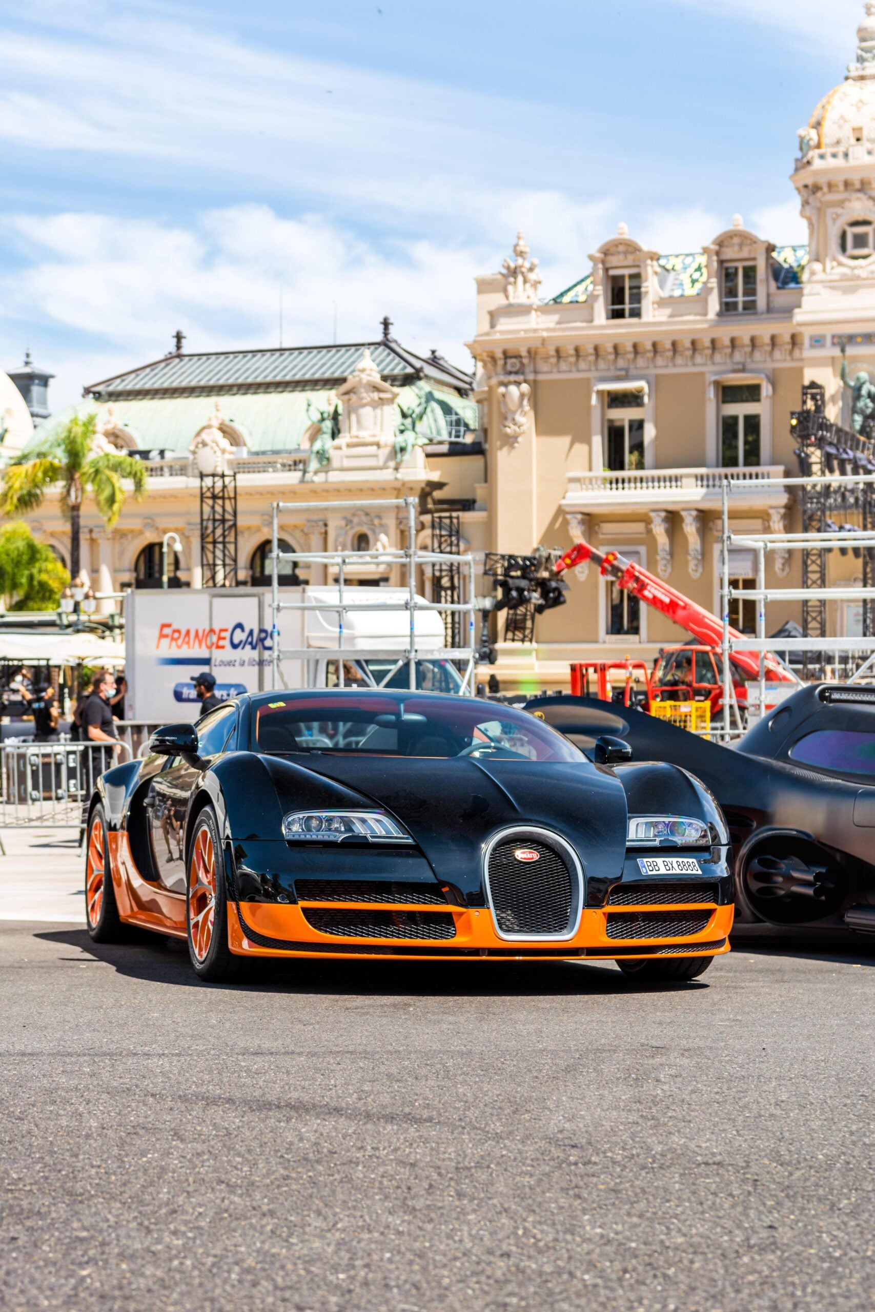 A black and orange Bugatti sports car is parked on a sunny street in front of ornate, historic European buildings, with people and another car in the background.