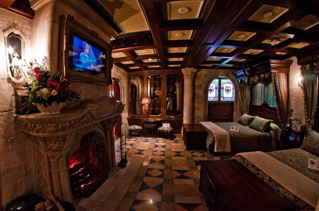 Lavishly decorated castle-themed bedroom with ornate fireplace, stained glass windows, carved stone walls, and two elegant beds. A TV is mounted above the fireplace, and the room features patterned floors and rich wooden ceiling panels.