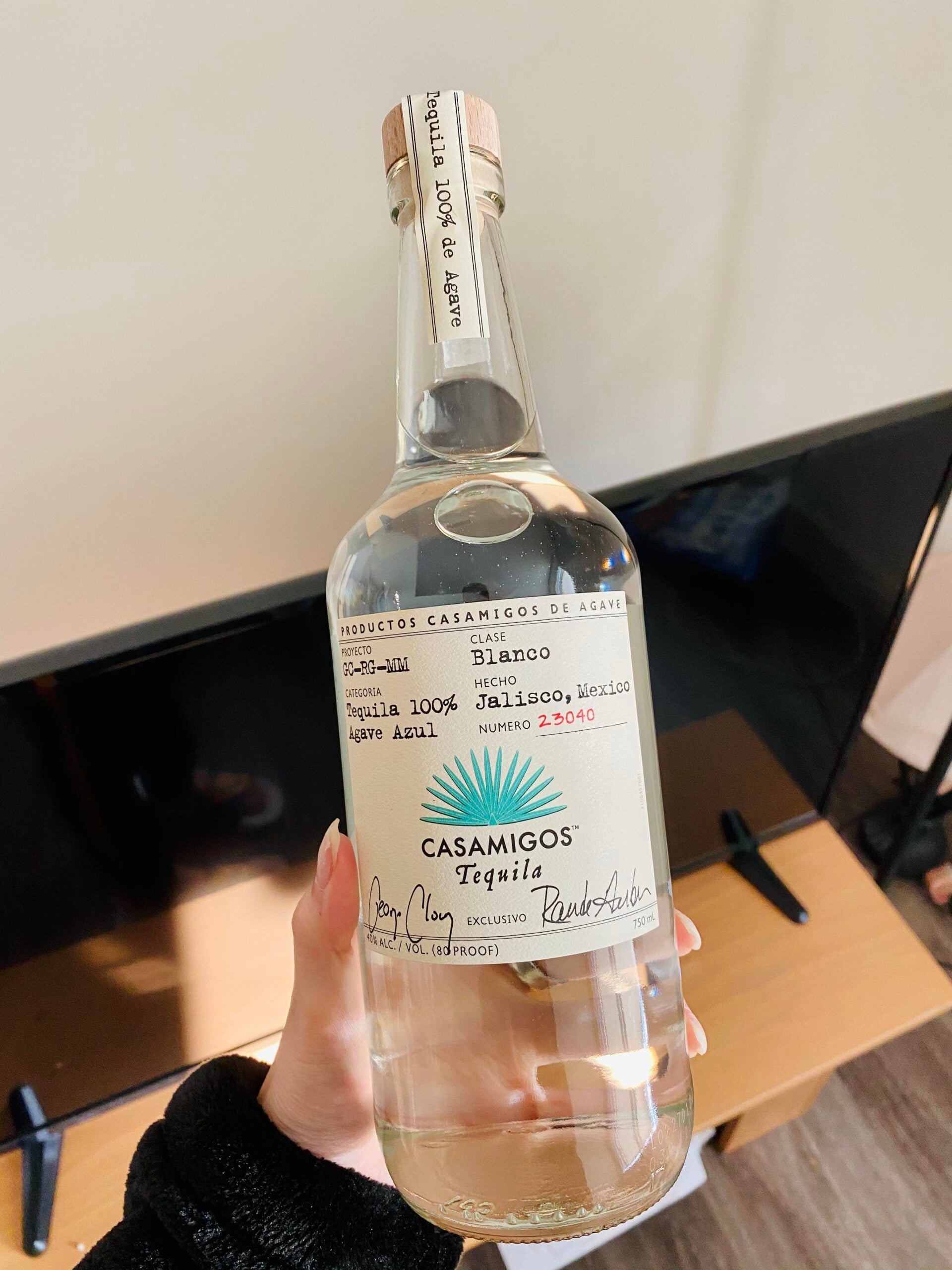 A hand holds a clear bottle of Casamigos Tequila Blanco with a white label in front of a TV and wooden table. The label shows signatures and handwritten numbering.