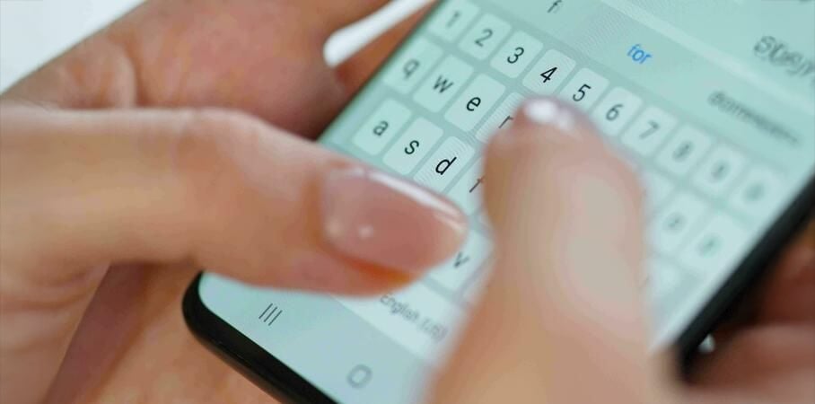 Close-up of a person’s hands typing on a smartphone’s touchscreen keyboard, with the focus on their fingers and part of the device's screen showing text input.