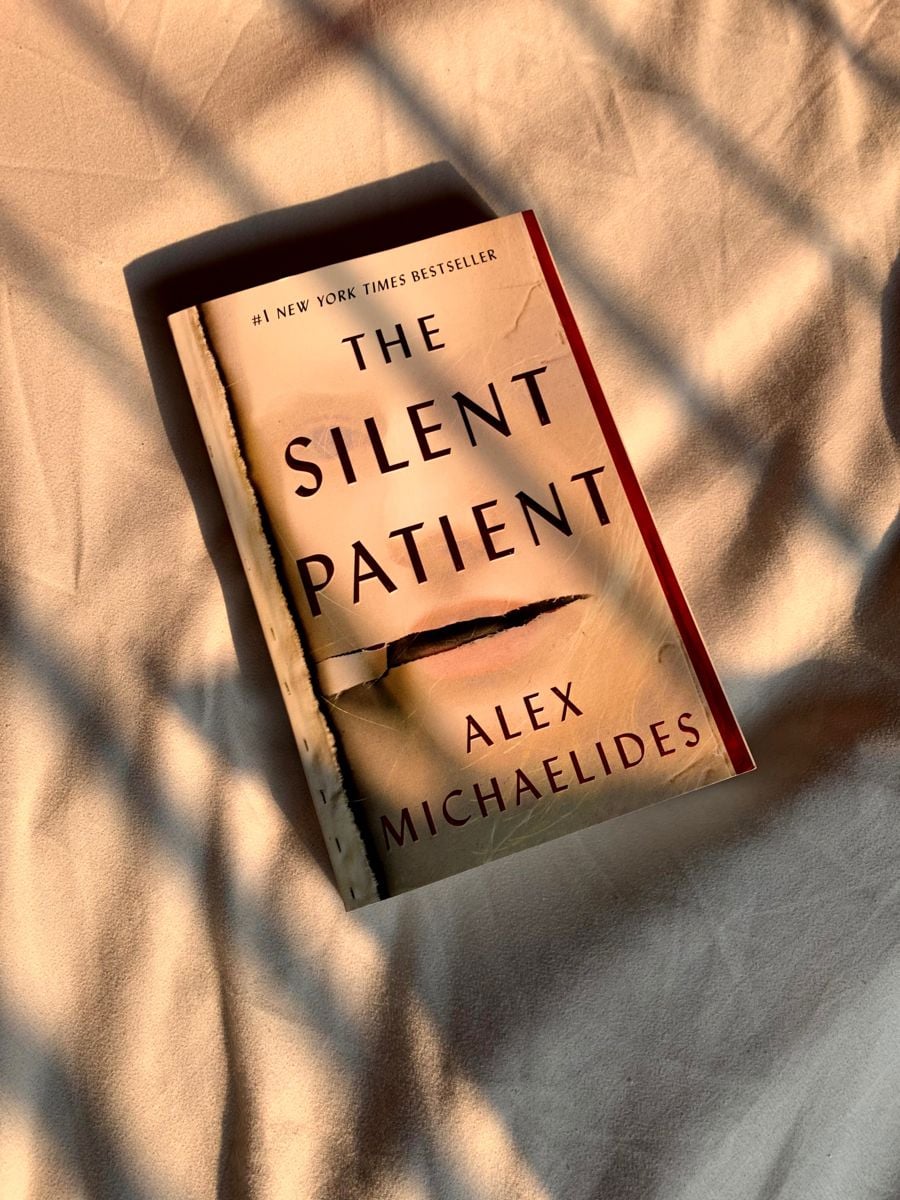 A copy of the book "The Silent Patient" by Alex Michaelides lies on a beige surface, with shadows of window blinds falling across the cover.