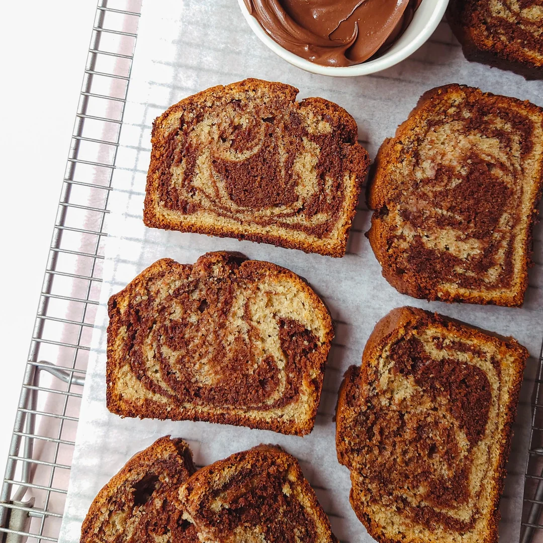 Slices of marble cake with chocolate and vanilla swirls are arranged on a cooling rack lined with parchment paper. A small bowl of chocolate spread sits nearby in the top right corner.