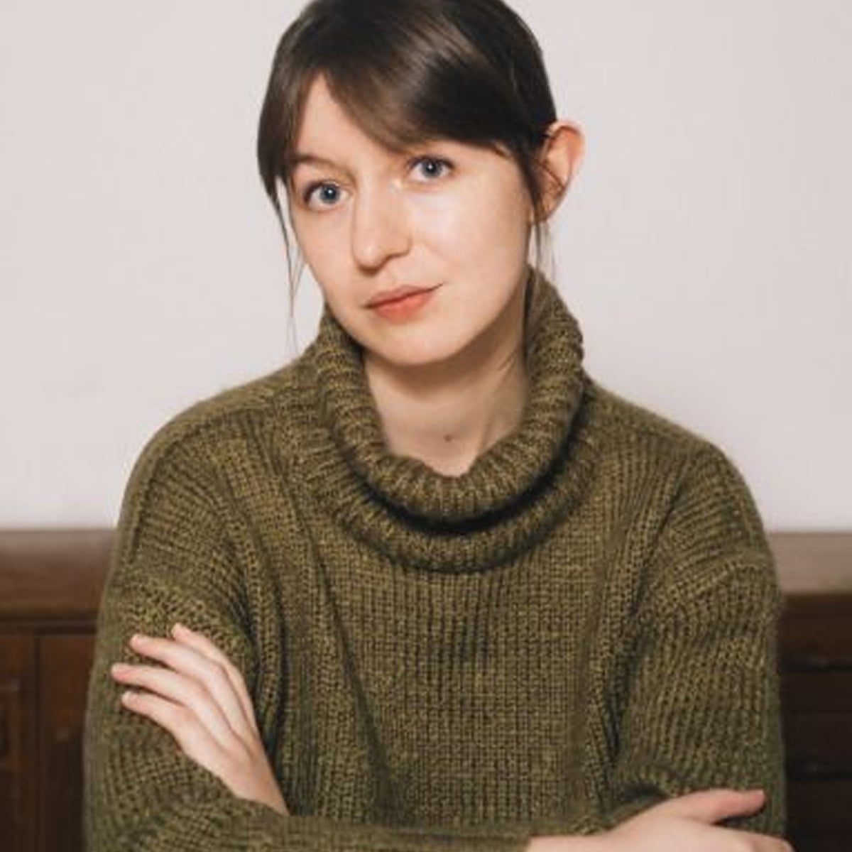 A young woman with light skin and straight brown hair wearing a chunky olive green turtleneck sweater sits with her arms crossed, looking at the camera with a neutral expression. The background is plain and indoor.