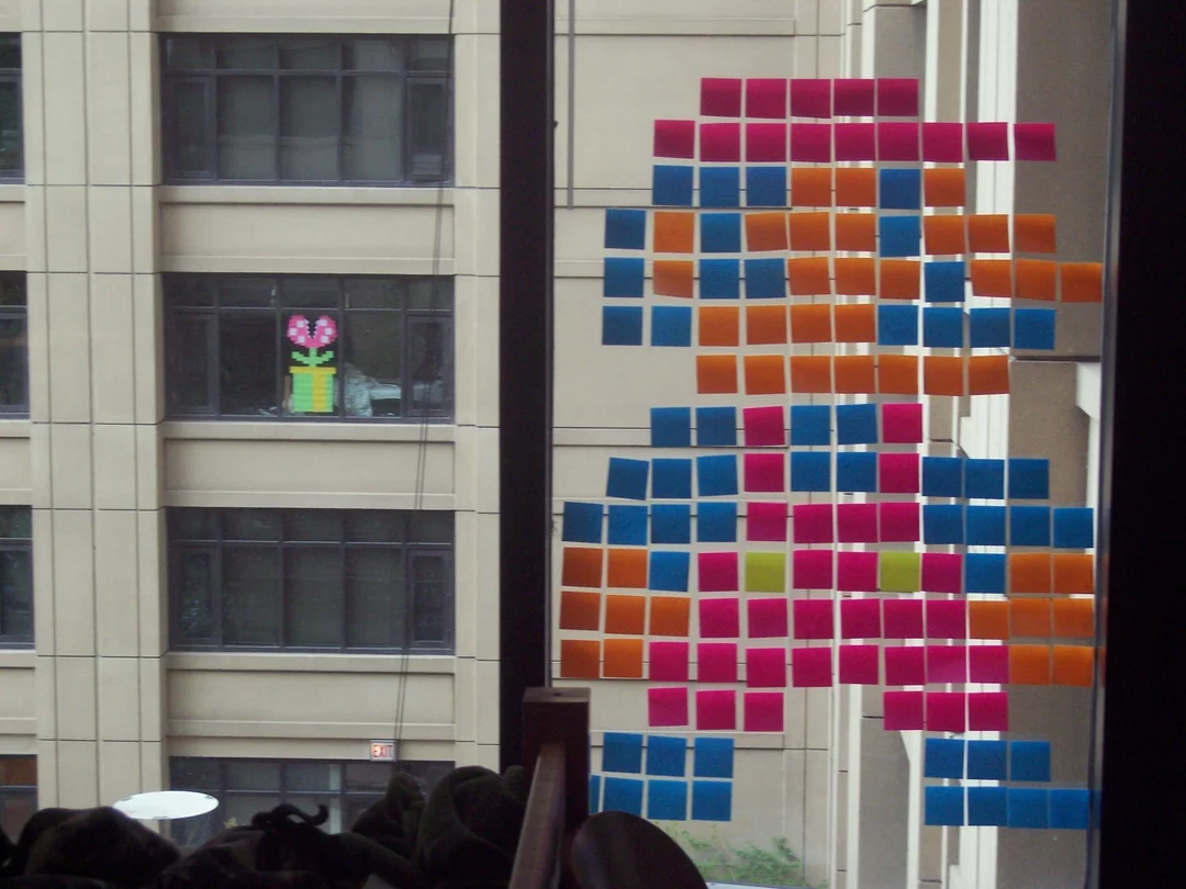 A window decorated with colorful sticky notes arranged to form an 8-bit Mario character; outside, another window displays a Piranha Plant from Mario, also made with sticky notes.
