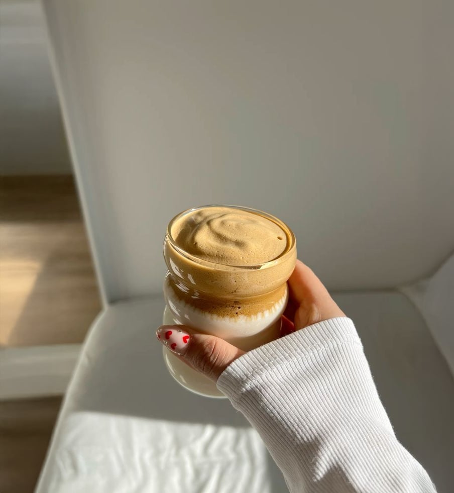 A hand with red heart nail art holds a glass of whipped dalgona coffee, featuring a frothy coffee layer over milk, in a sunlit room with white furniture.