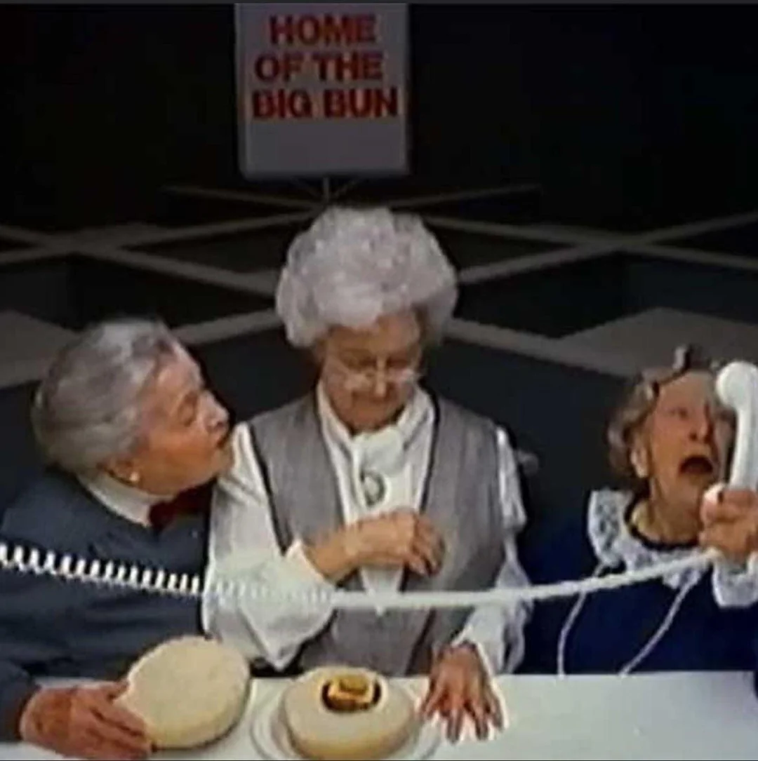 Three elderly women examine a large hamburger bun with a tiny burger patty inside. One woman shouts into a phone, while a sign in the background reads "Home of the Big Bun.