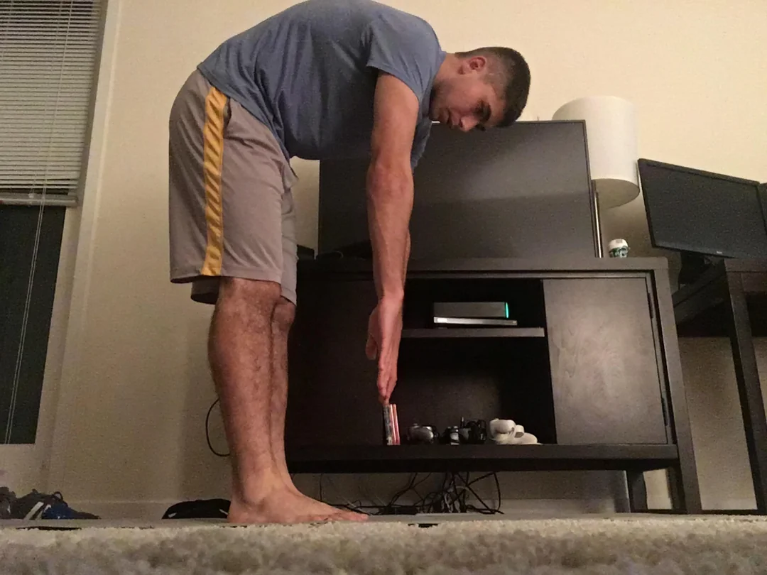 A man in athletic shorts and a t-shirt stands indoors on carpet, bending forward with straight knees, reaching toward the floor but not touching it. Electronic devices and cords are visible on the dark wooden furniture behind him.