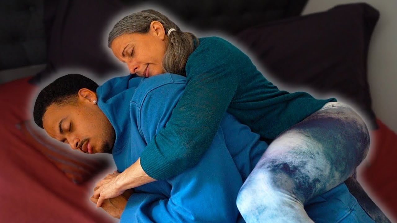A woman with gray hair hugs a man from behind while both sit on a bed. The man appears calm with his eyes closed, and the woman rests her head gently on his shoulder.