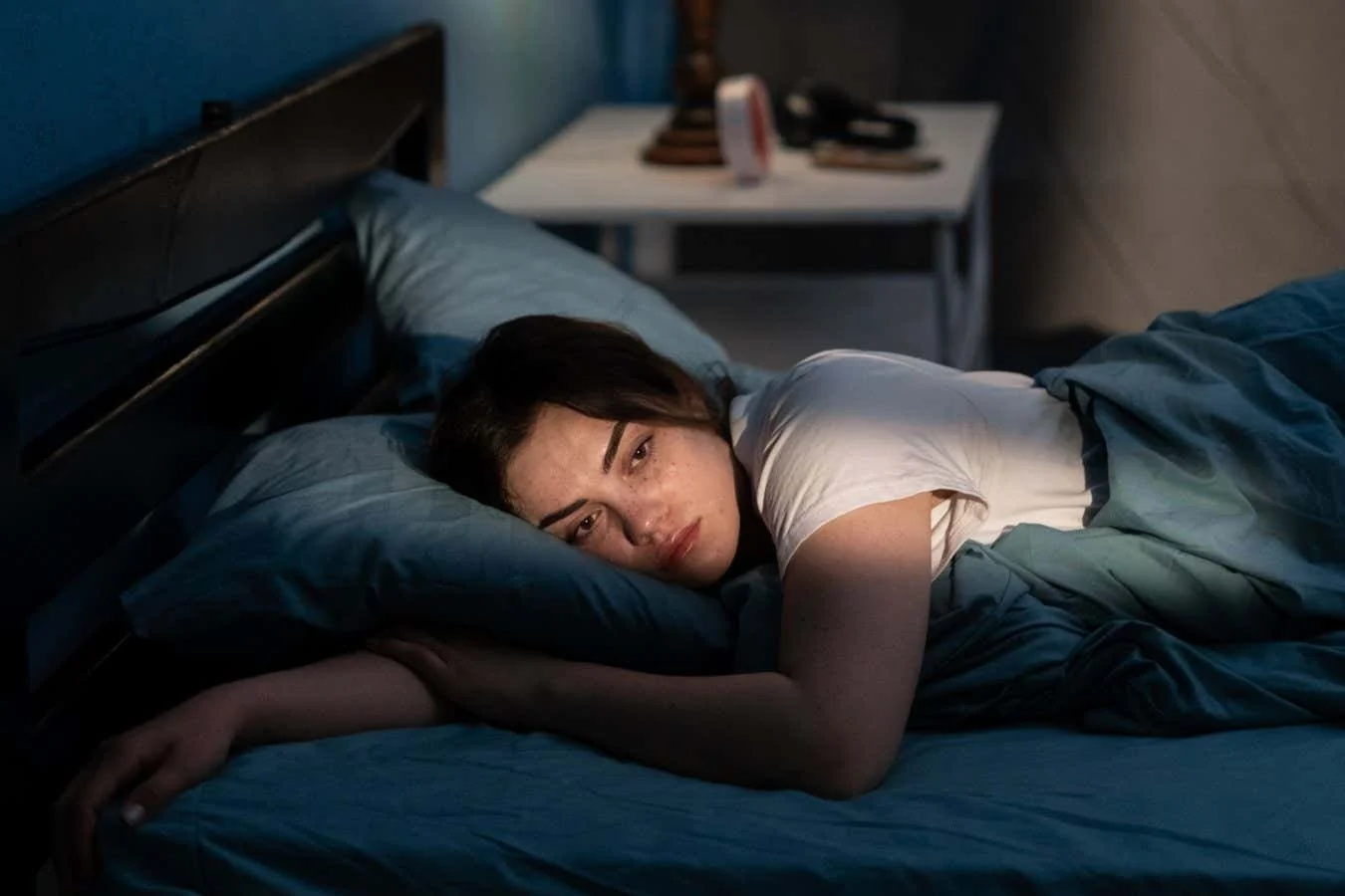 A woman lies in bed on her side, looking sad with tears on her face. She is partially covered by a teal blanket, resting her head on a pillow, with soft light casting a shadow across the room.