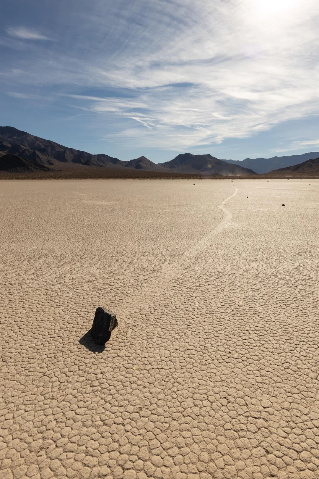A large rock rests on a dry, cracked desert floor under a partly cloudy sky, with a long track behind it suggesting the rock has moved across the landscape. Distant mountains line the horizon.