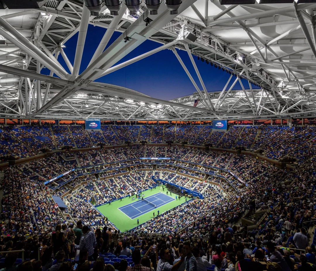A crowded tennis stadium under a partially open roof at dusk, with bright lights illuminating the court where a match is taking place. Spectators fill the stands, and the sky is deep blue above the structure.