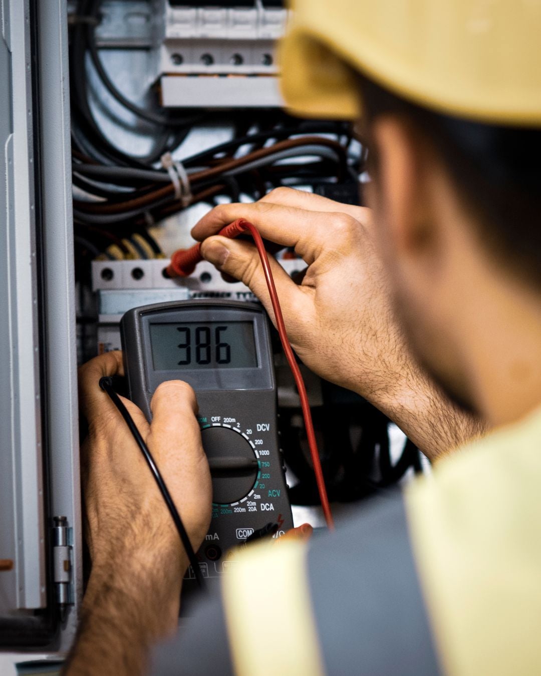 A person wearing a yellow hard hat uses a multimeter to measure voltage inside an electrical panel, with the display reading 386. The person holds test probes to wires and wears a reflective safety vest.