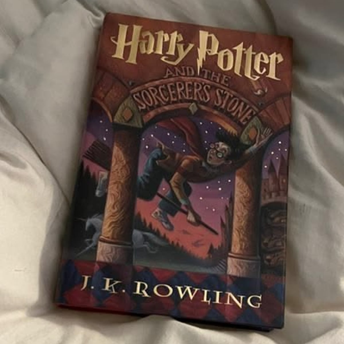 A copy of "Harry Potter and the Sorcerer's Stone" by J.K. Rowling rests on a light-colored, rumpled fabric surface. The book cover shows Harry flying on a broomstick, reaching for a golden ball.