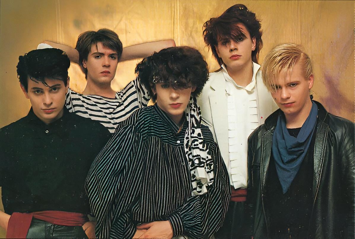 Five young men pose together against a gold background, wearing 1980s-style clothing such as striped shirts, patterned tops, and jackets. Their hairstyles are voluminous and styled, reflecting 1980s fashion trends.