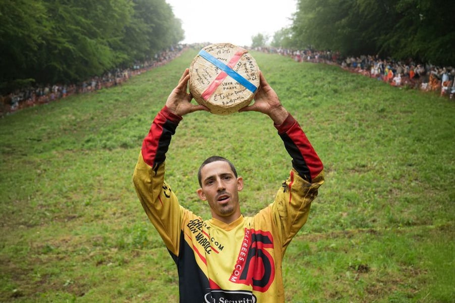 A man in a yellow and red jersey holds a large, round cheese above his head at the bottom of a steep, grassy hill, with a crowd lining both sides of the slope in the background.