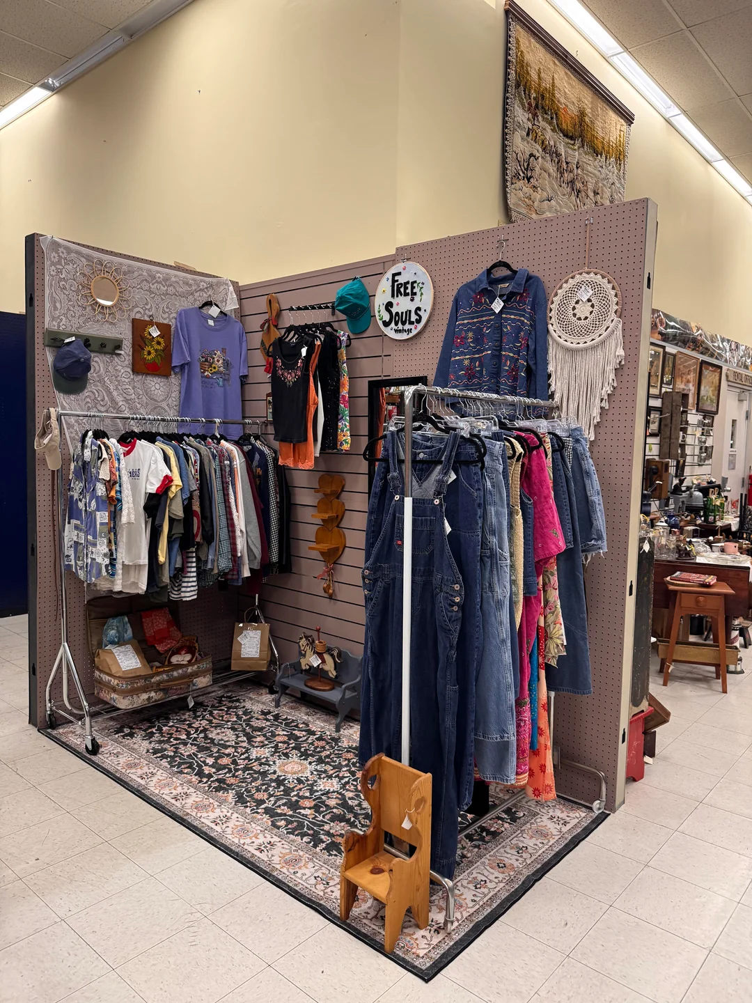 A vintage clothing booth features colorful shirts, jackets, and overalls on racks and hangers, with accessories and decor like hats, a dreamcatcher, and a “Free Souls” sign displayed on the walls.