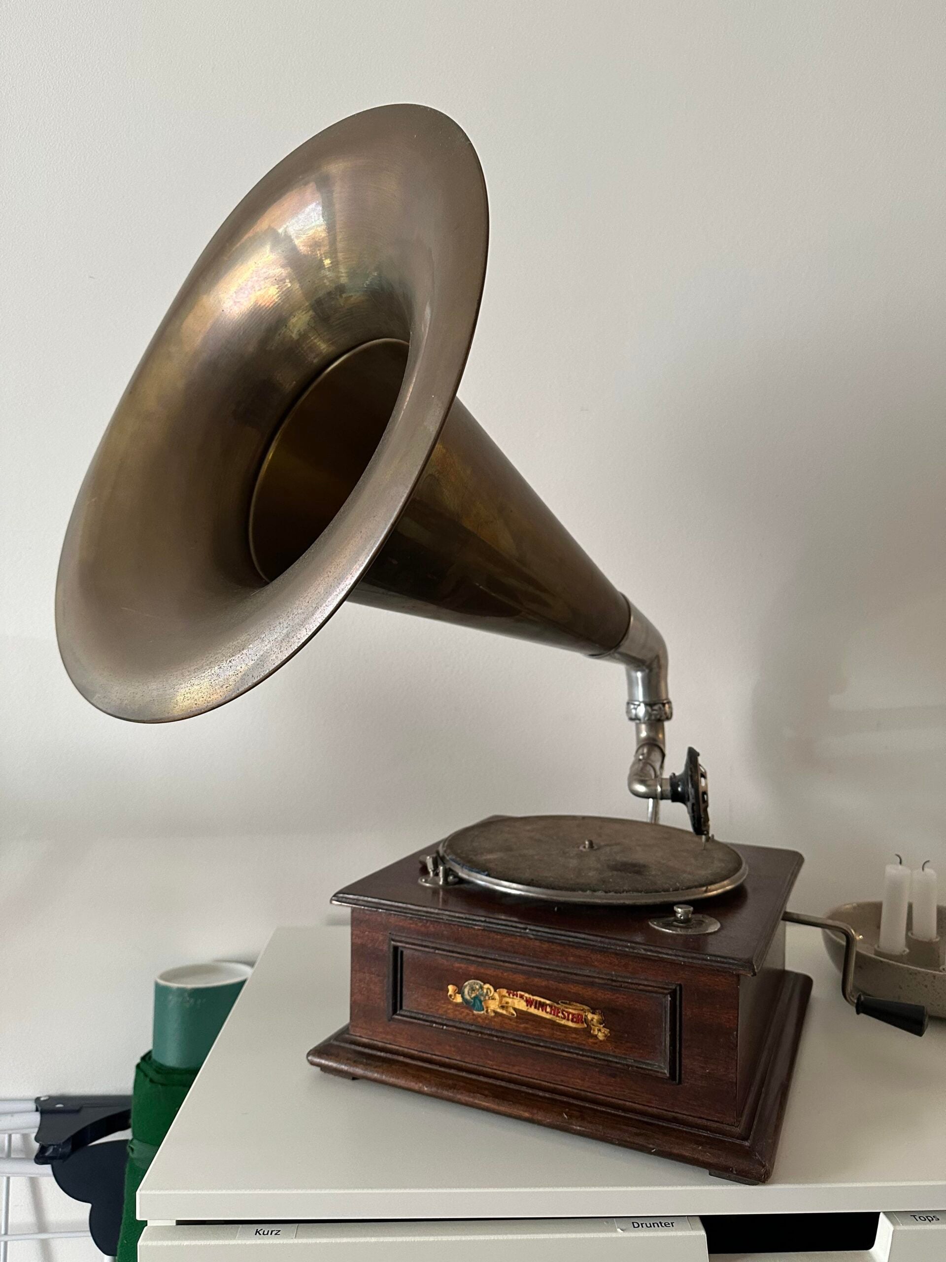 An antique gramophone with a large brass horn and a wooden base sits on a white surface against a plain wall. The turntable and decorative details are visible on the front.