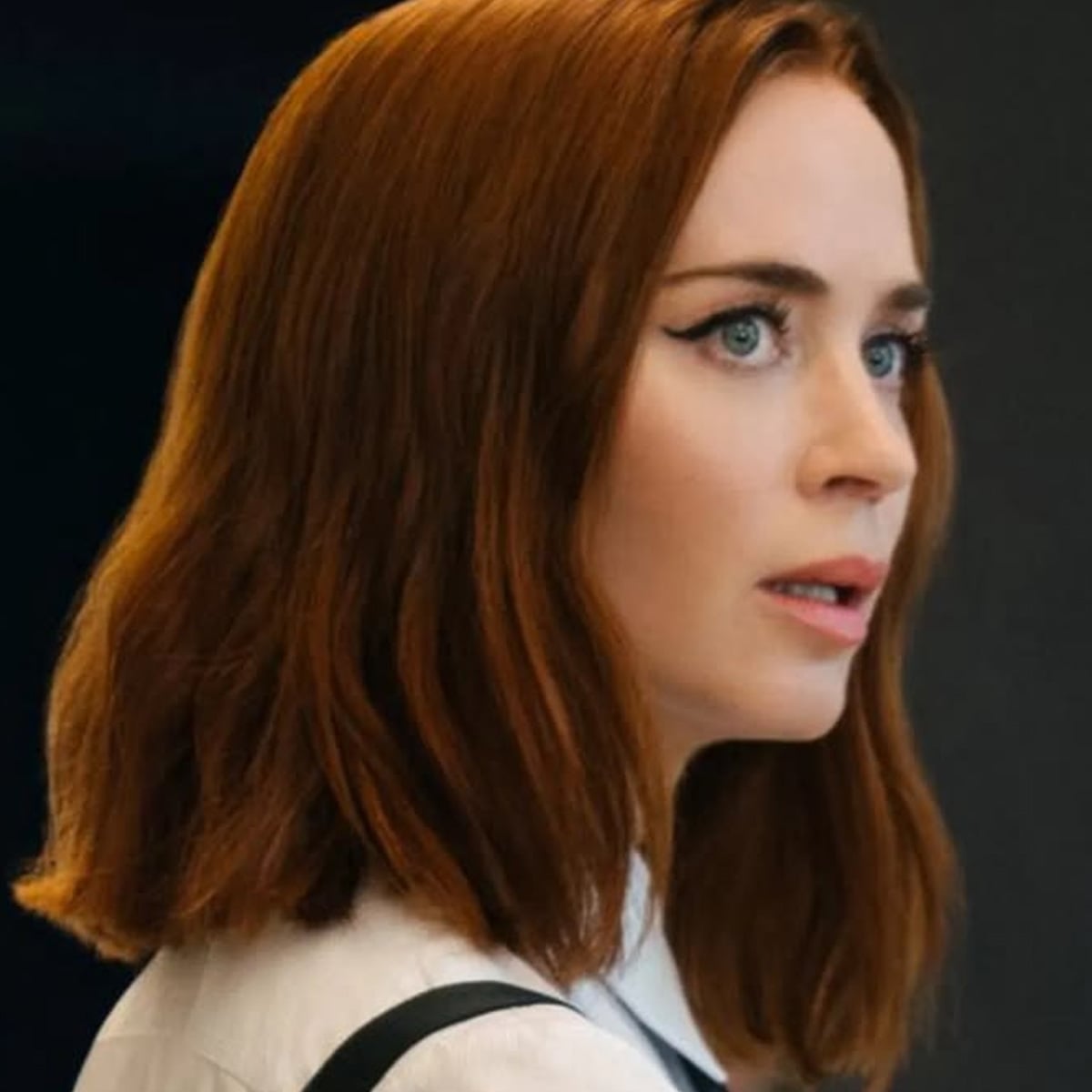 A woman with shoulder-length auburn hair and blue eyes, wearing a white collared shirt, looks intently to the side against a dark background.