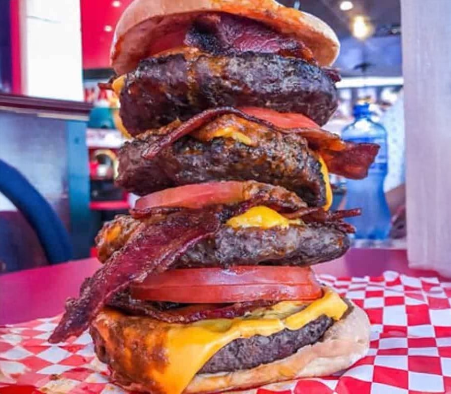 A towering burger with four beef patties, layers of melted cheese, bacon, and tomato slices on a bun, served on red checkered paper in a diner setting.
