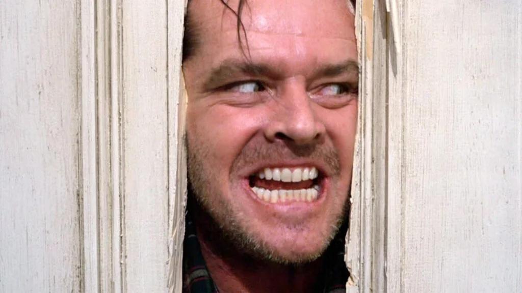 A man with a wild expression on his face peers through a broken door, his teeth bared in a menacing grin and his face partially framed by splintered wood.