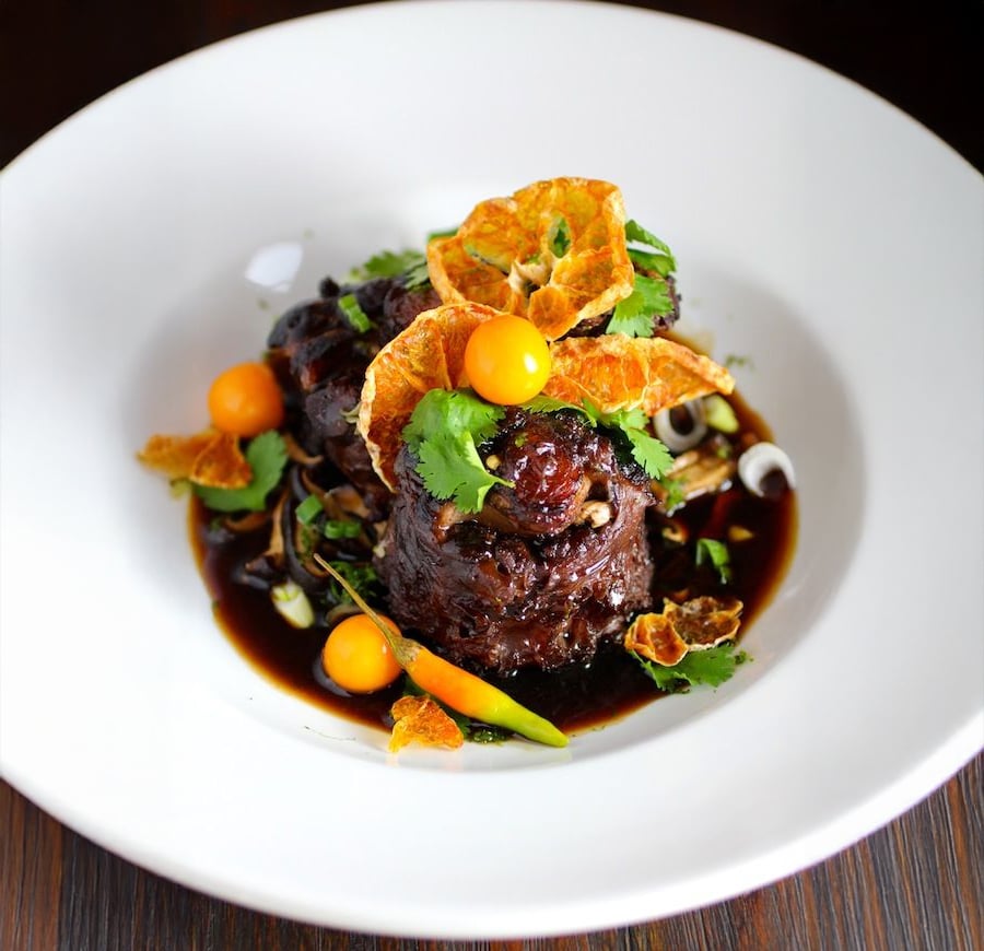 A gourmet dish featuring braised meat in dark sauce, garnished with fresh cilantro, crispy orange slices, golden berries, green vegetables, and herbs, served on a white plate against a dark background.
