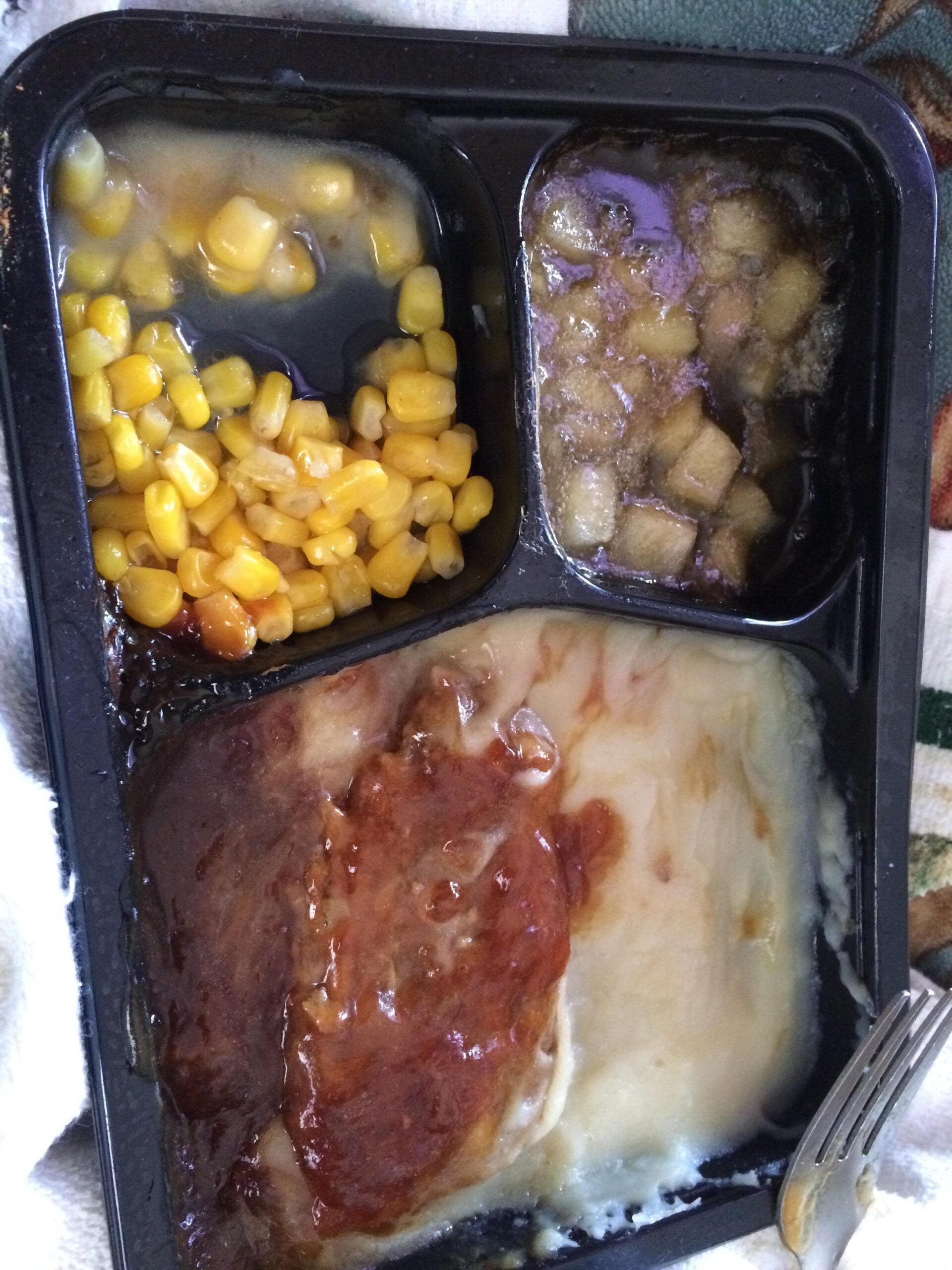 A TV dinner with corn in sauce, diced apples in syrup, and sliced beef with gravy over mashed potatoes in a black tray, with part of a fork visible at the bottom right.