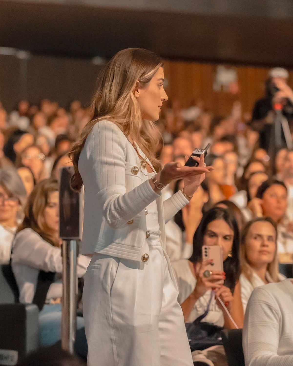 A woman in a white suit speaks to a large seated audience at an indoor event. She gestures with one hand while holding a small device in the other. Many attendees watch and some hold up phones to record.