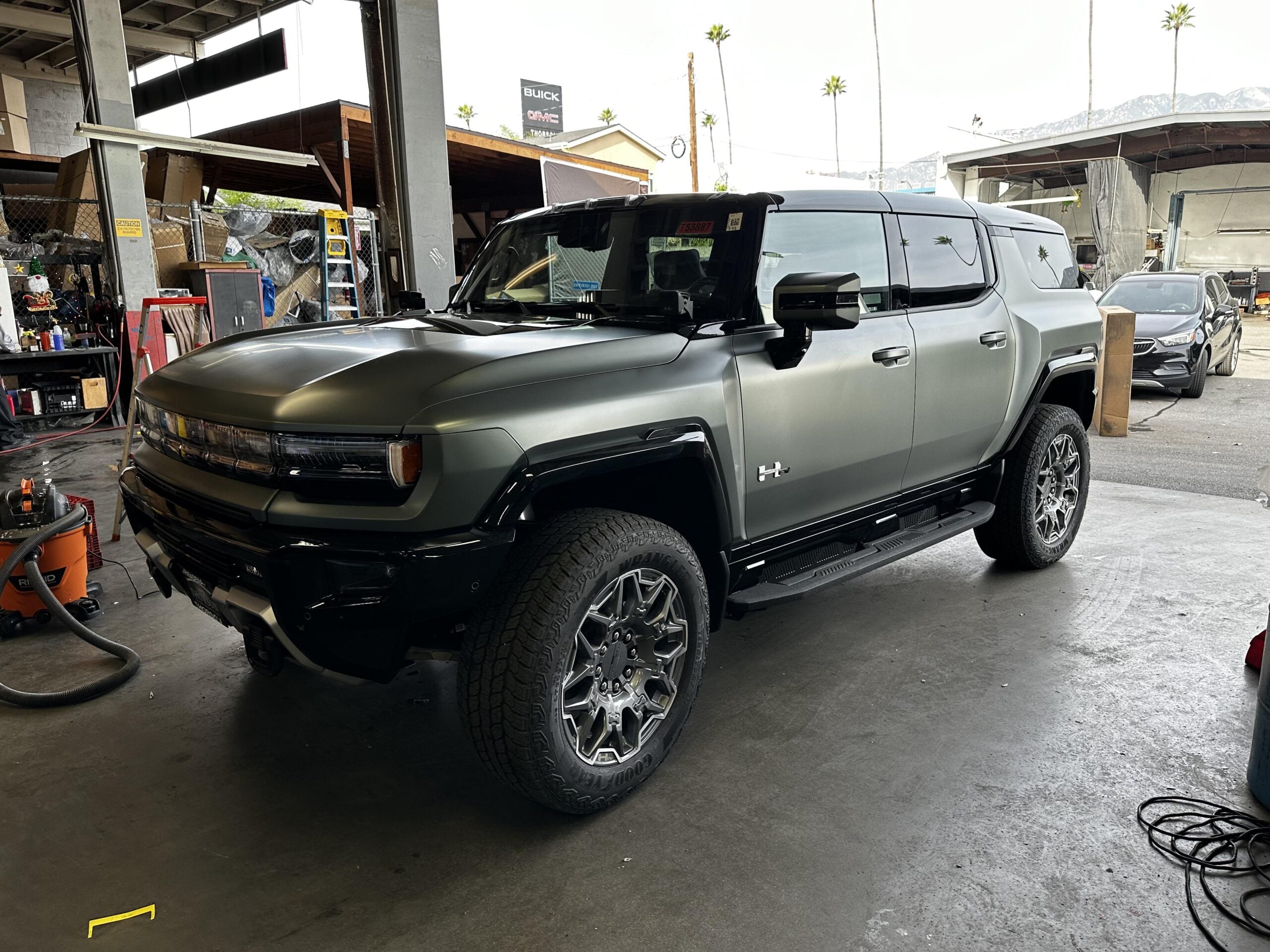 A matte green GMC Hummer EV SUV is parked inside an industrial garage, with equipment and boxes scattered around and mountains visible in the background.