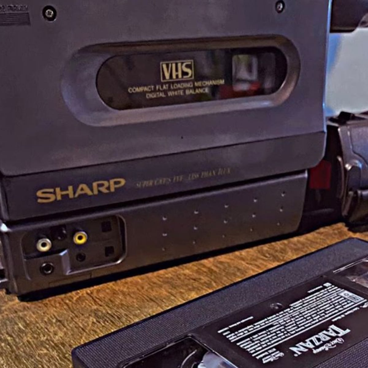 A close-up of a vintage Sharp VHS player with visible input ports and a VHS tape labeled "TITAN" lying on a wooden surface in front of it.