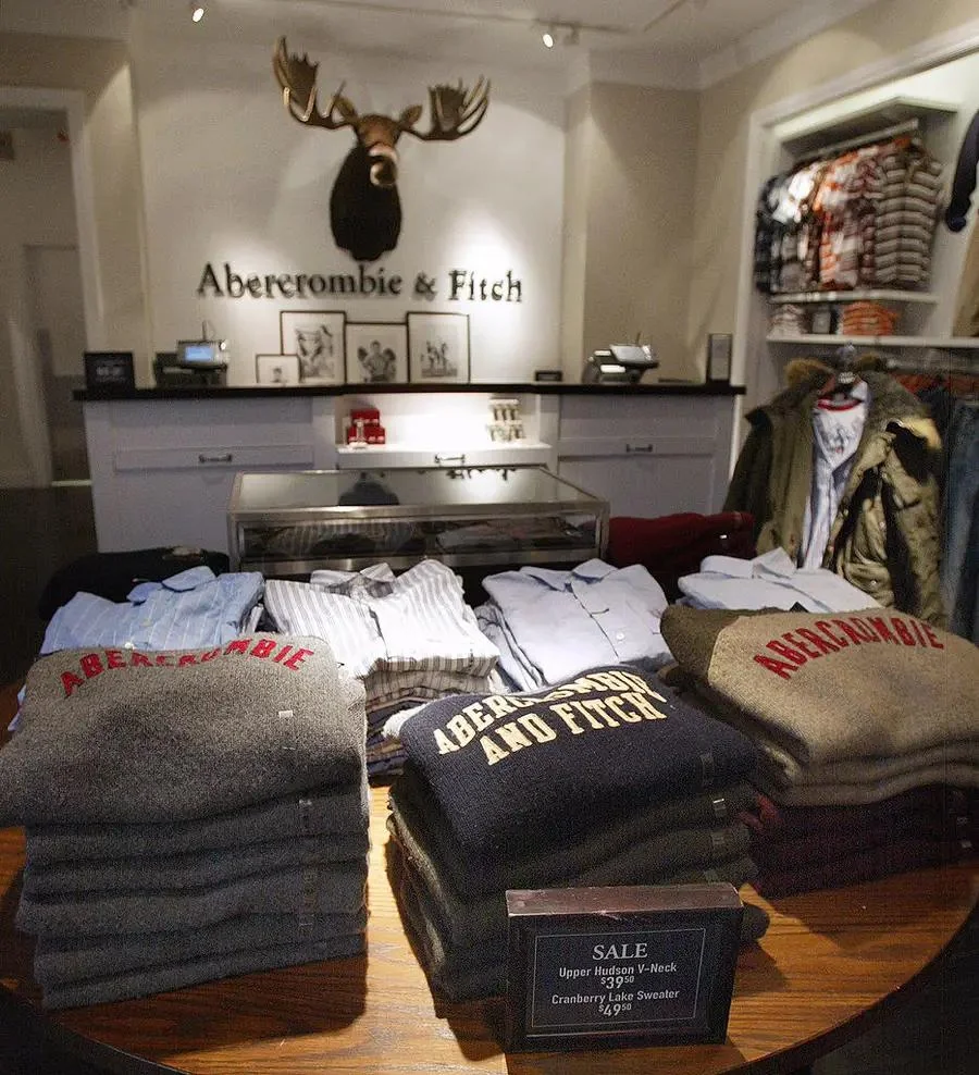 A display inside an Abercrombie & Fitch store shows neatly folded sweaters in gray tones, some with the brand name on them. A moose head and the store logo are mounted on the wall above the display.