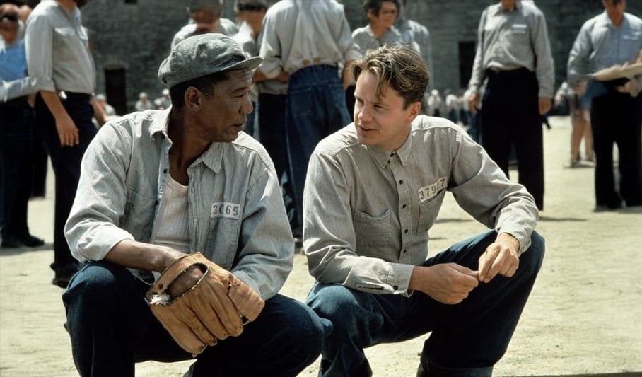 Two men in prison uniforms crouch on a dirt yard, talking closely. One holds a baseball glove. Other inmates in similar uniforms stand around in the background.