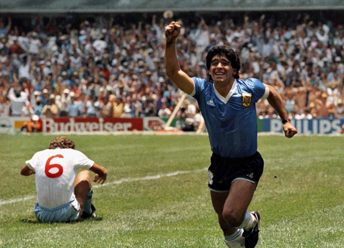 An Argentine soccer player celebrates a goal with his arm raised, while an English player wearing number 6 sits on the grass in disappointment. A large crowd fills the stadium in the background.