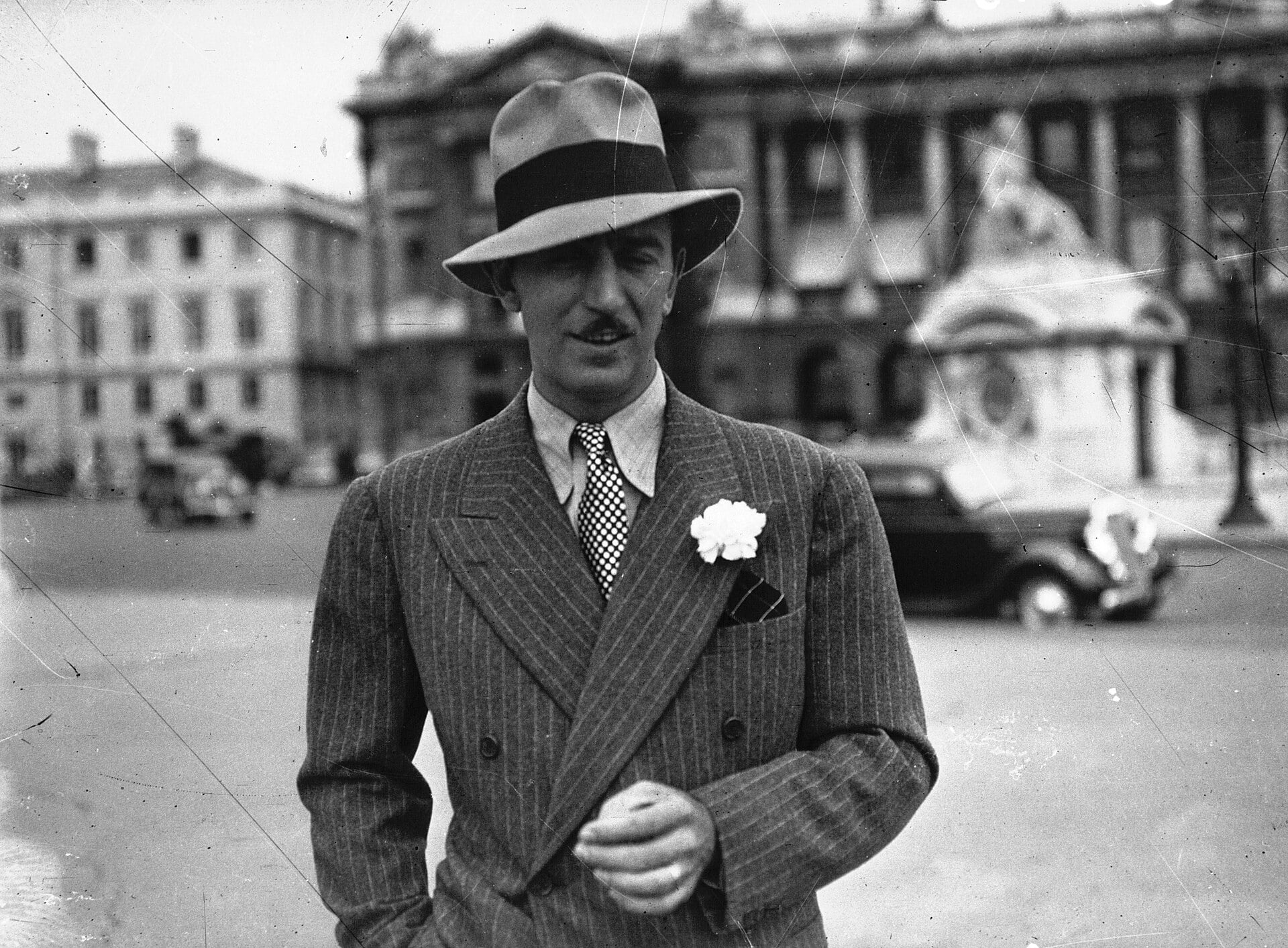 A man in a pinstripe suit, tie, and wide-brimmed fedora stands outdoors in front of historic buildings and vintage cars. He has a white flower in his lapel and looks toward the camera.