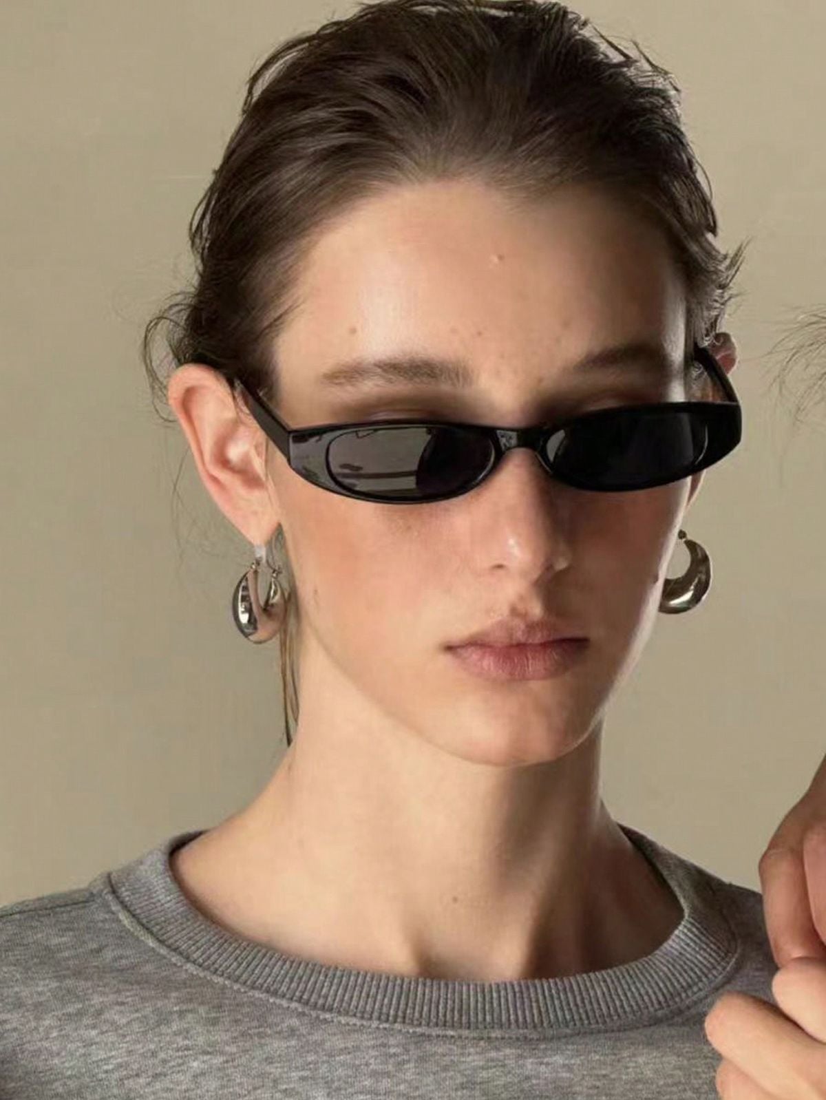 A person with light skin and brown hair pulled back wears narrow black sunglasses, silver hoop earrings, and a gray crew-neck top, standing against a plain beige background.