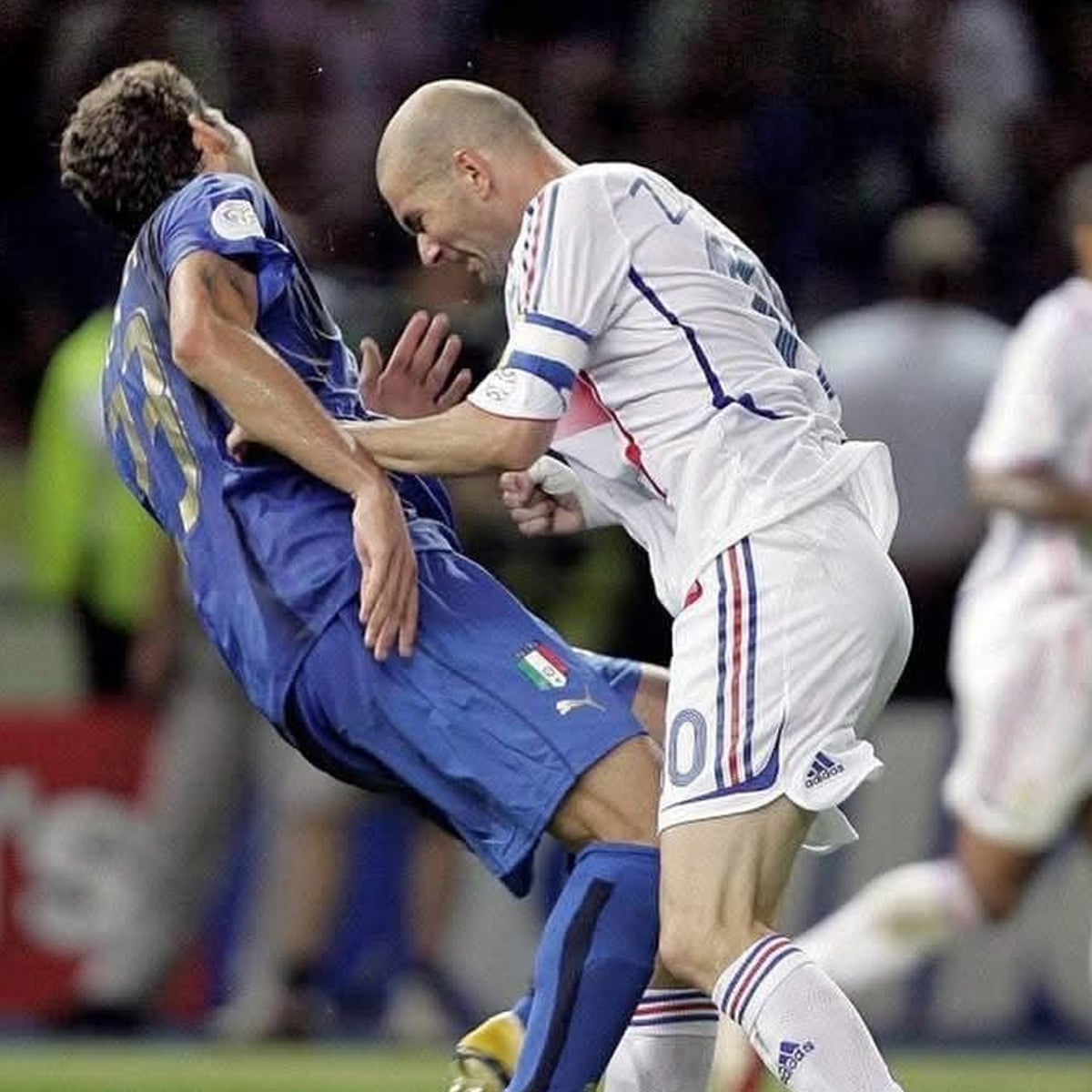 A soccer player in a white jersey headbutts an opponent in a blue jersey during a match, causing the blue-clad player to bend backward in reaction. Both are on a grassy field with blurred players and spectators in the background.