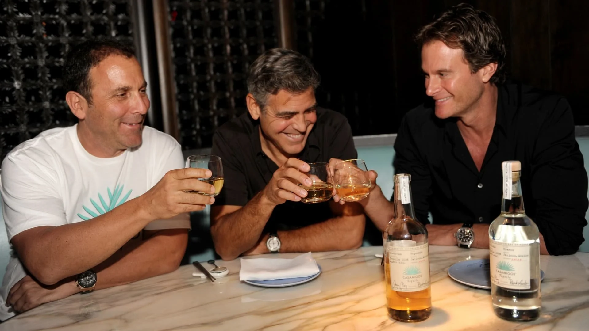 Three men sit at a marble table, smiling and toasting with glasses of liquor. There are two bottles on the table, and the setting appears to be a restaurant or bar with dim lighting and wine storage in the background.