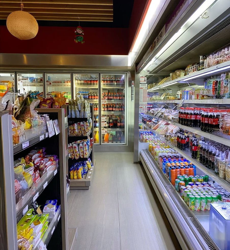 A brightly lit grocery store aisle with shelves stocked with snacks, drinks, and packaged foods. Refrigerated beverages are on the right and at the back, with chips and other snacks on display to the left.