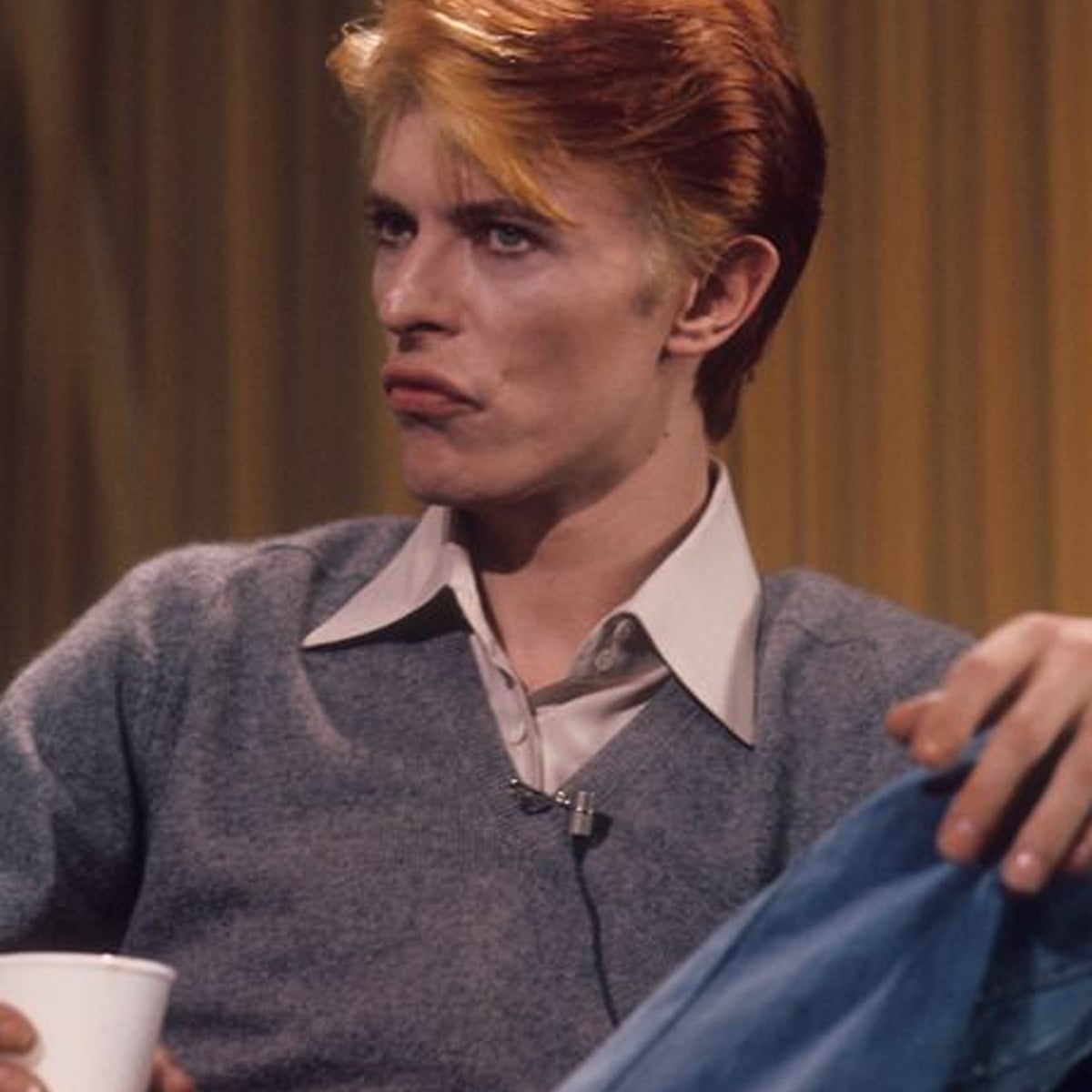 A person with light skin and bright orange-blonde hair sits casually, holding a white cup. They wear a gray sweater over a collared shirt and blue jeans, with a serious expression against a gold curtain backdrop.