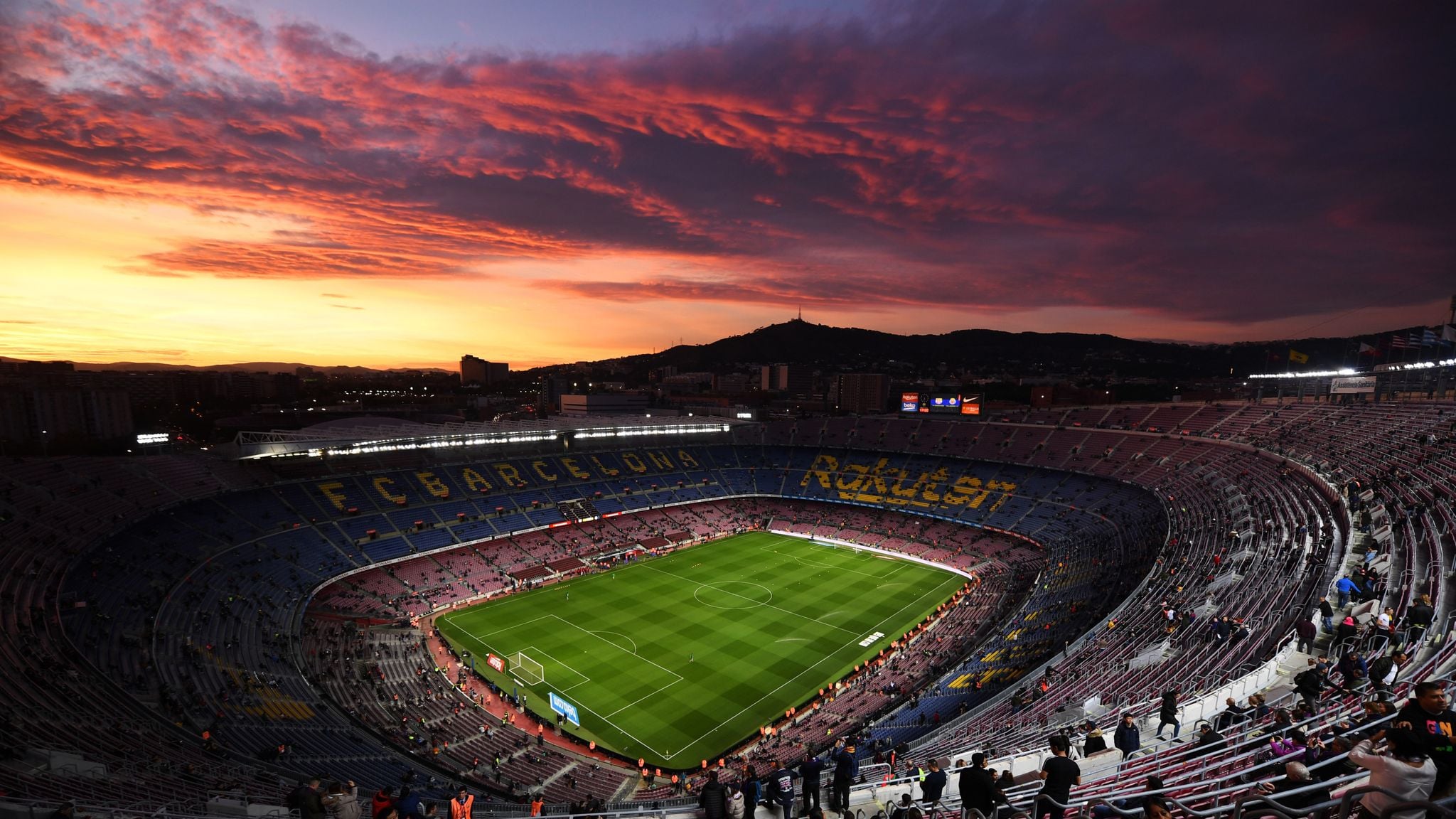 A panoramic view of FC Barcelona’s Camp Nou stadium at sunset, with a glowing sky, empty seats, and the field illuminated for an upcoming match. The city and hills are visible in the background.