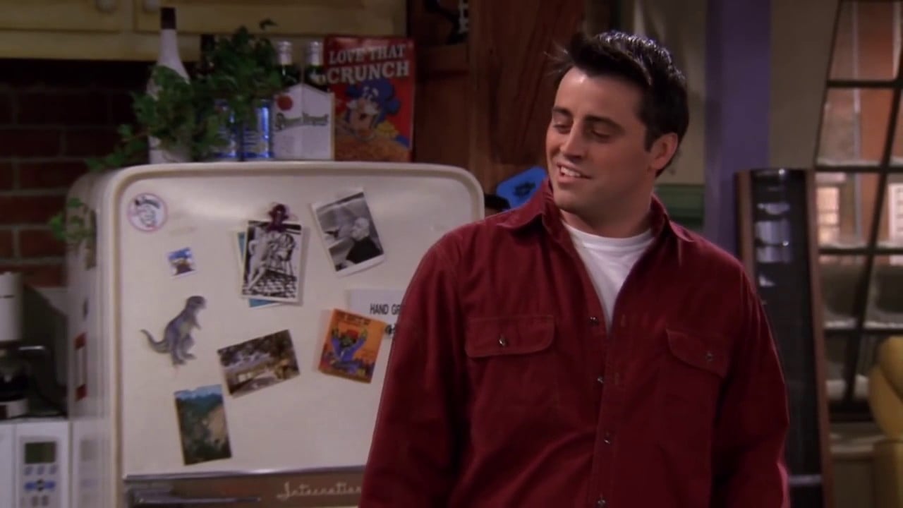 A man in a red shirt stands smiling in a cozy kitchen. Behind him, a white fridge is decorated with photos, magnets, and postcards. A box of cereal sits on top of the fridge.