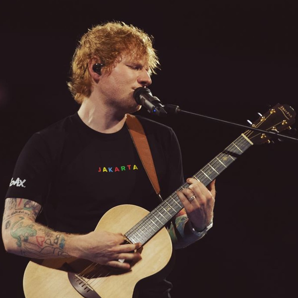 A man with reddish hair plays an acoustic guitar and sings into a microphone. He wears a black "Jakarta" t-shirt and has colorful tattoos on his arms. The background is dark, focusing attention on him.