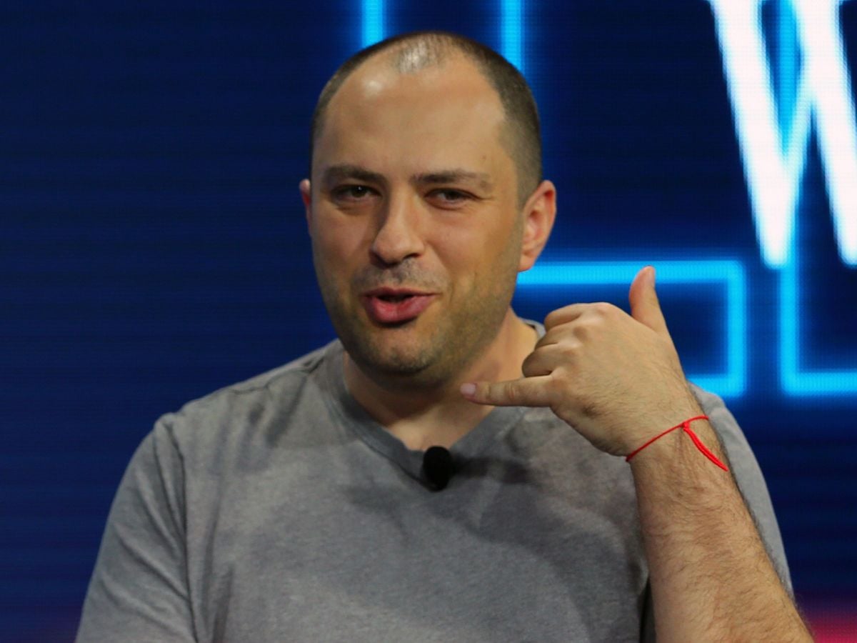 A man with short hair wearing a gray t-shirt makes a phone gesture with his right hand. He has a red string bracelet on his wrist and is speaking, with a blue background behind him.