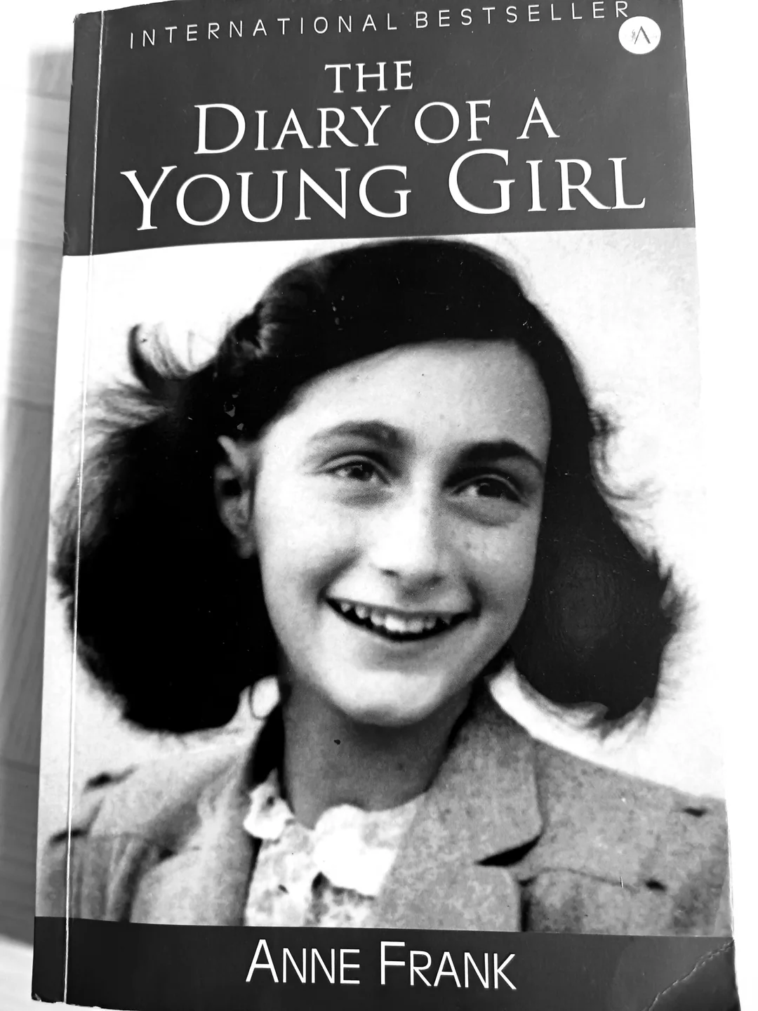 Black and white book cover of "The Diary of a Young Girl" by Anne Frank, featuring a smiling young girl with dark hair wearing a light-colored jacket and white shirt. The top says "International Bestseller.