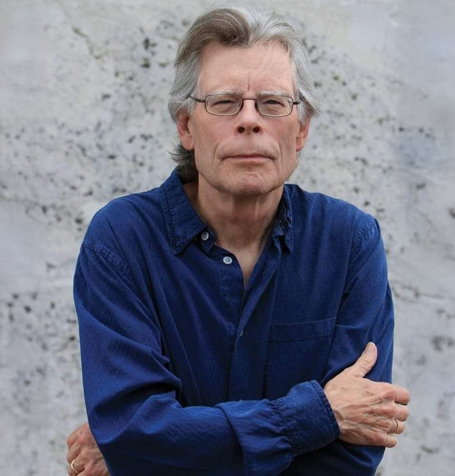 An older man with gray hair and glasses, wearing a blue button-up shirt, stands with his arms crossed in front of a light gray textured wall.
