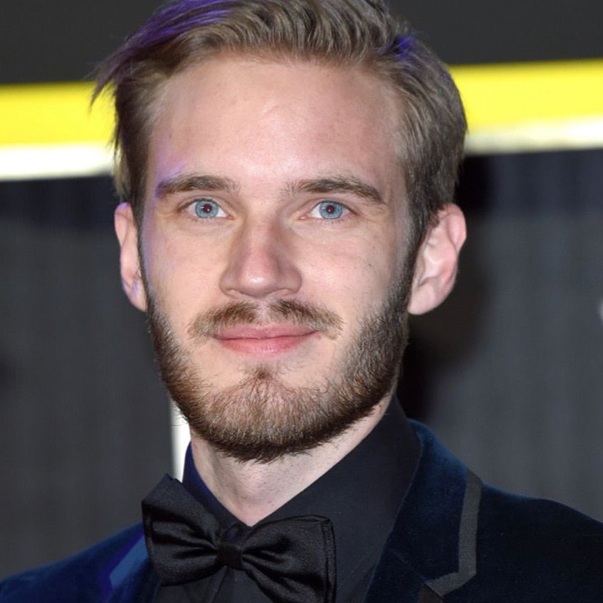A man with light skin, blue eyes, and light brown hair and beard, wearing a dark velvet suit, black shirt, and black bow tie, smiles slightly while looking at the camera. The background is out of focus.