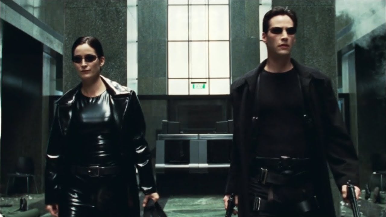 Two people in black leather outfits and sunglasses walk confidently through a modern, glass-walled lobby, each holding a gun. The scene has a dramatic, action-oriented atmosphere.