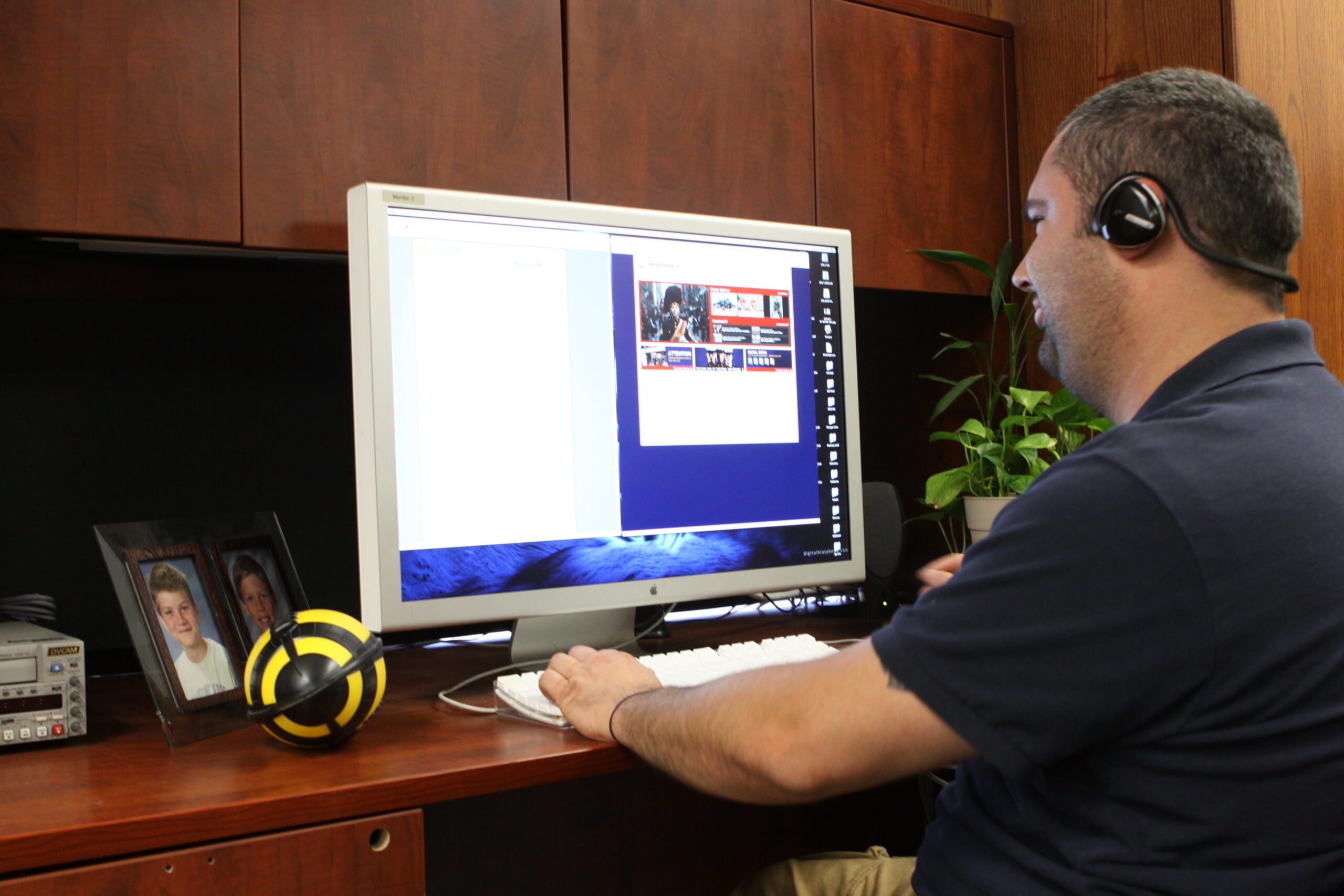 A man wearing a headset sits at a desk using a computer. There is a framed photo, a toy resembling a bee, and a potted plant on the desk. The computer screen shows multiple open windows.