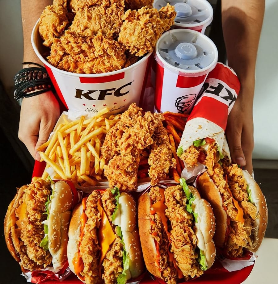 A person holds a large tray of KFC food, including buckets of fried chicken, fries, three chicken burgers with lettuce, fried chicken pieces, two soft drinks, and a chicken wrap.